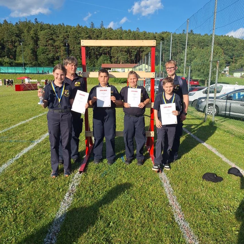 Eine Gruppe von fünf Jugendlichen, die Uniformen und Medaillen tragen, steht auf einem Sportplatz, hält Urkunden in den Händen und posiert für ein Foto. Hinter ihnen steht eine rote Konstruktion, und mehrere Autos sind im Hintergrund geparkt.