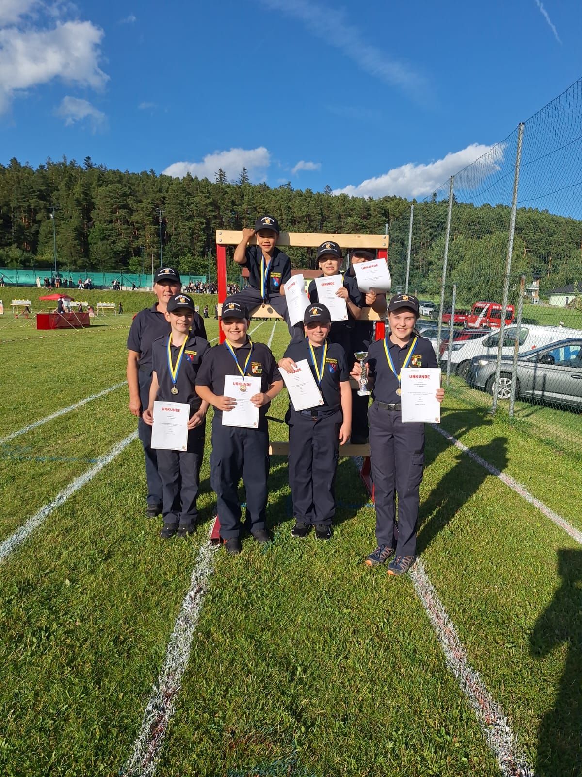 Eine Gruppe junger Menschen in Uniformen und Mützen, die Zertifikate halten, steht auf einem Feld. Dahinter sind ein Zaun, Bäume und geparkte Autos zu sehen.