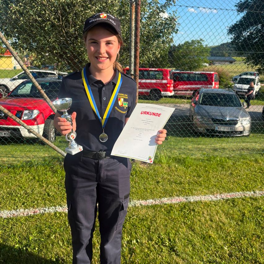 Eine junge Frau in Uniform hält ein Zertifikat und eine Trophäe und lächelt. Hinter ihr steht ein Zaun mit geparkten Autos und einem roten Bus. Bäume und ein Gebäude sind im Hintergrund.