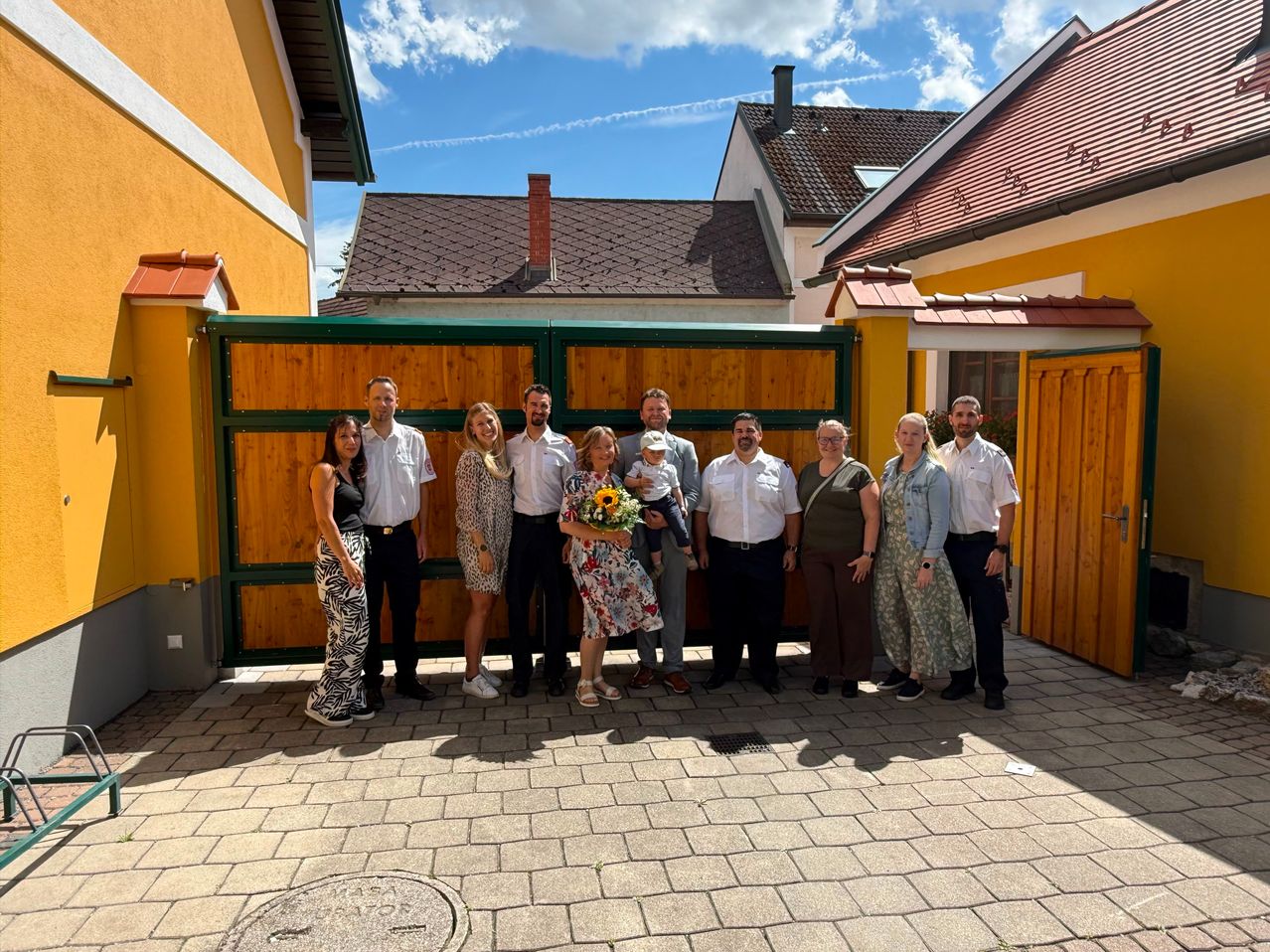 Eine Gruppe von Menschen steht vor einem Haus. Sie lächeln und posieren für ein Foto. Die Frau in der Mitte hält ein Baby und einen Blumenstrauß.