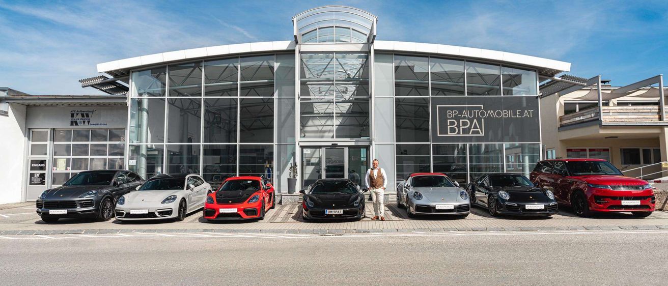Ein Mann steht vor einem BP-Automobile.at-Händler. Mehrere Sportwagen sind draußen geparkt, darunter ein weißer Porsche, ein roter Porsche und ein schwarzer Porsche.
