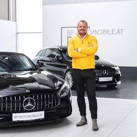 Ein Mann in einem gelben Pullover steht neben zwei schwarzen Mercedes-Benz Autos in einer Ausstellung. Hinter ihm ist ein großes weißes Schild mit dem BP-Automobile.at Logo.