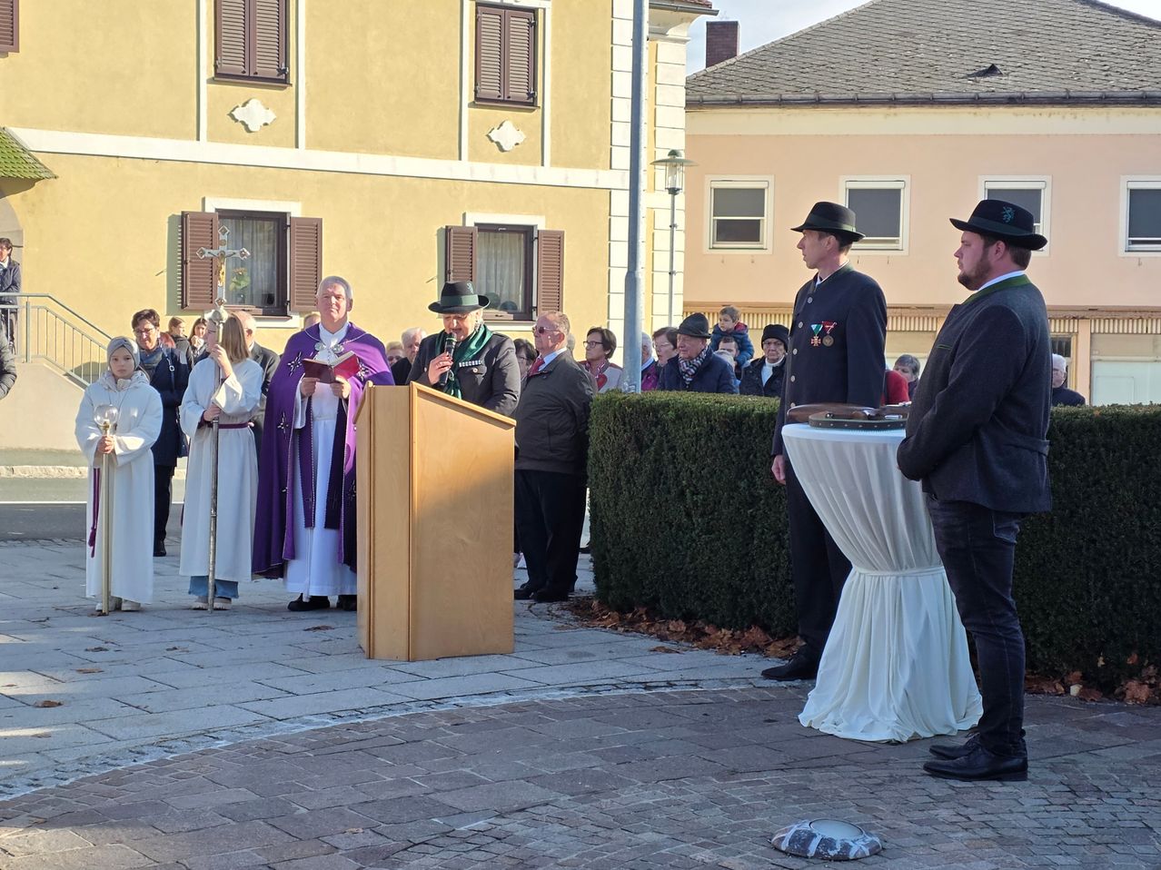 A solemn event is taking place outdoors. Several individuals are present, including a man with a microphone at a podium. A priest reads from a book, and others observe.