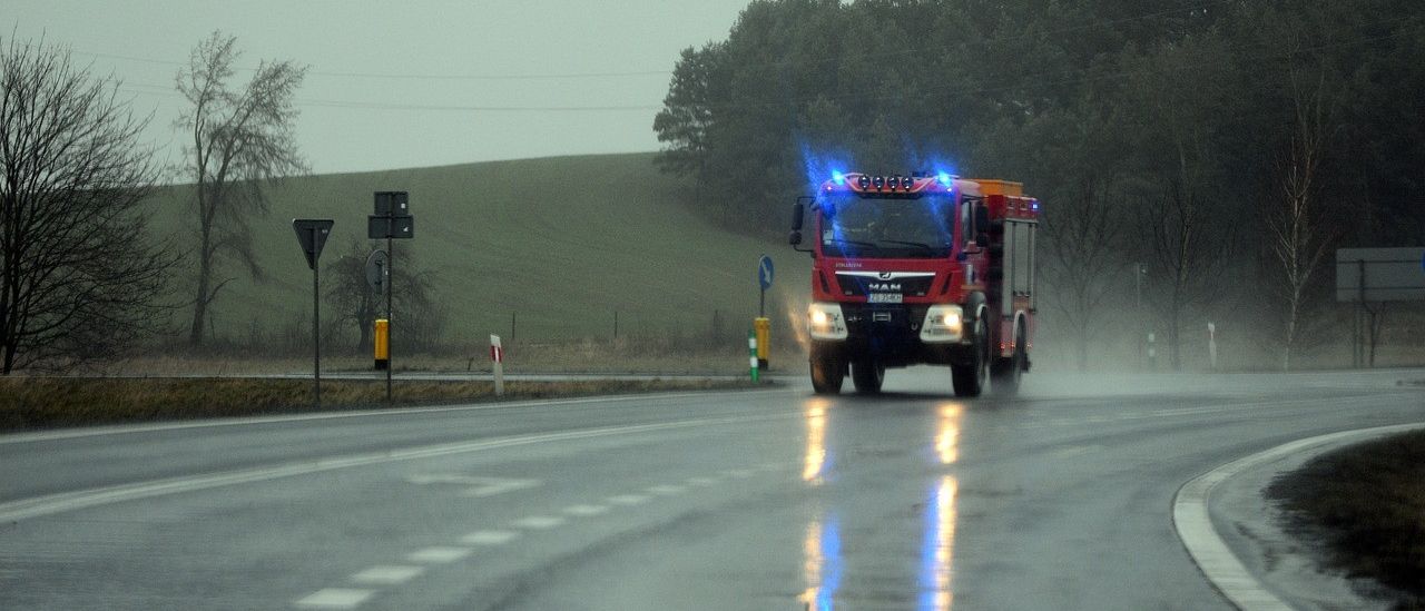 Ein roter Feuerwehrwagen mit blauen Lichtern fährt auf einer nassen Straße. Der Himmel ist bedeckt. Bäume und ein Hügel sind im Hintergrund.