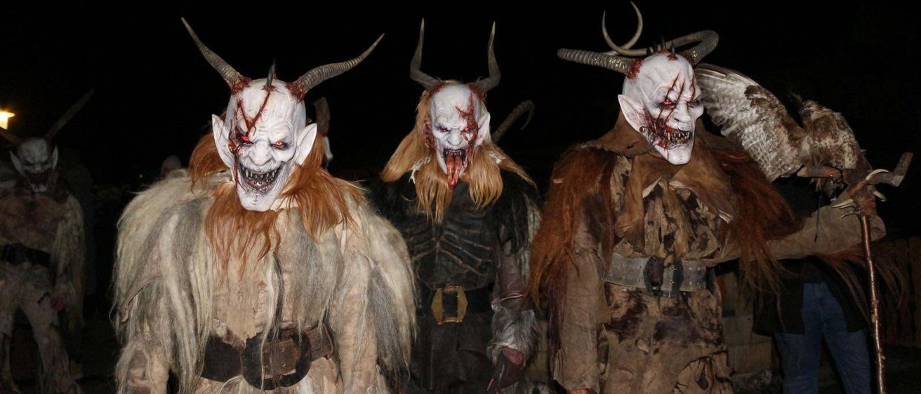 Mehrere Personen in aufwendigen blutbespritzten Hörnermasken stehen in einem dunklen Bereich. Sie sind in zerlumpte Kleidung gekleidet, eine hält einen Zweig in der Hand.