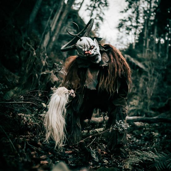 Eine Person mit einer gruseligen Maske mit Hörnern und langen Haaren steht in einem dunklen Wald.