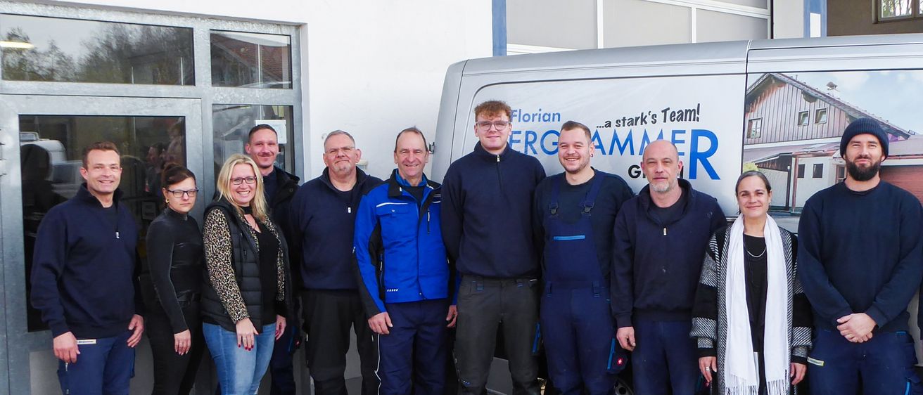 Eine Gruppe von Menschen in blauen Uniformen steht vor einem Gebäude mit einem Schild, das 'Ghammer GmbH' besagt. Das Gebäude hat eine Glastür und Fenster. Ein Van mit dem Text 'Ghammer' ist vor dem Gebäude geparkt.