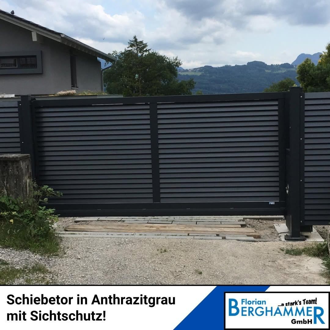 Ein modernes schwarzes Schiebetor mit Lamellen ist vor einem Haus mit Blick auf einen Berg im Hintergrund installiert.
