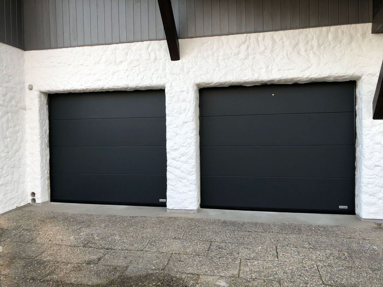 Zwei schwarze Garagentore mit einer strukturierten weißen Wand im Hintergrund, ein Tor hat ein kleines silbernes Logo in der unteren linken Ecke.
