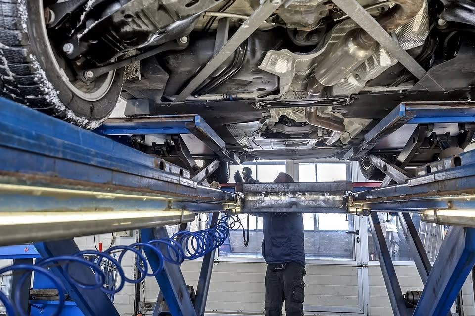 Ein Mechaniker arbeitet in einer Werkstatt unter einem Auto auf einer Hebebühne. Er trägt eine blaue Jacke und Hose. Es sind noch zwei weitere Personen an dem Auto tätig.