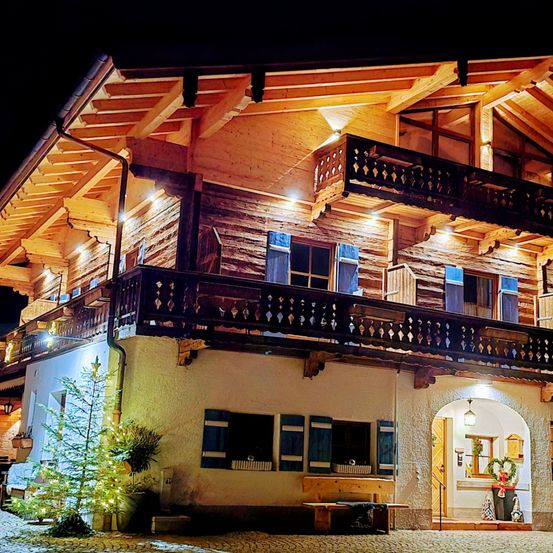 Ein warm beleuchtetes Holzchalet mit mehreren Balkonen, blauen Fensterläden und einem beleuchteten Weihnachtsbaum davor. Der Eingang ist beleuchtet, mit einer Bank und einem Mülleimer in der Nähe.