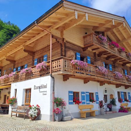 Ein Chalet mit hölzernen Balkonen und farbenfrohen Blumenkästen. Bänke und Topfpflanzen schmücken die Vorderseite unter einem klaren blauen Himmel.