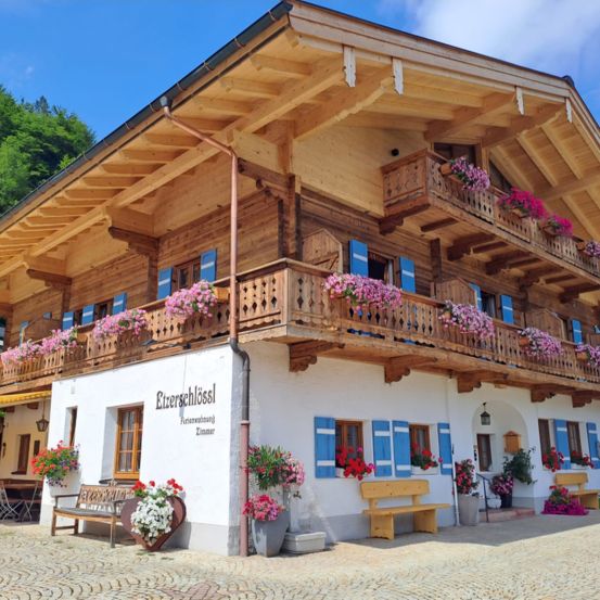 Ein Chalet mit hölzernen Balkonen und farbenfrohen Blumenkästen. Bänke und Topfpflanzen schmücken die Vorderseite unter einem klaren blauen Himmel.