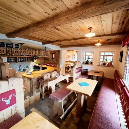 Das Innere eines rustikalen Restaurants mit Holzmöbeln, Holzbalkendecken und dekorativen Hirschkopf-Designs auf den Bankrücken. Der Küchenbereich ist sichtbar mit einer Bartheken und Regalen.