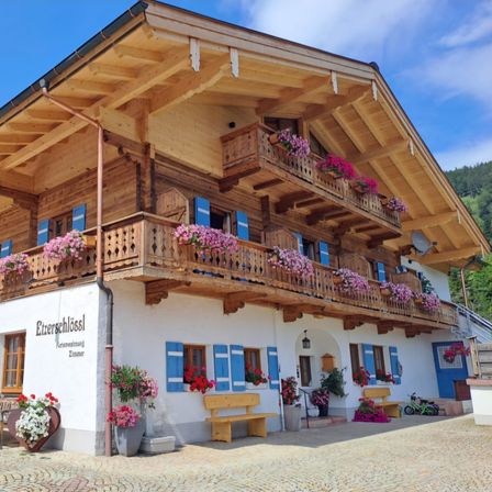 Ein Holzchalet mit mehreren Balkonen, die mit rosa Blumen geschmückt sind. Holzbänke und Topfpflanzen sind draußen. Das Gebäude hat ein Schild, das 'Eitzerschlossl' steht.