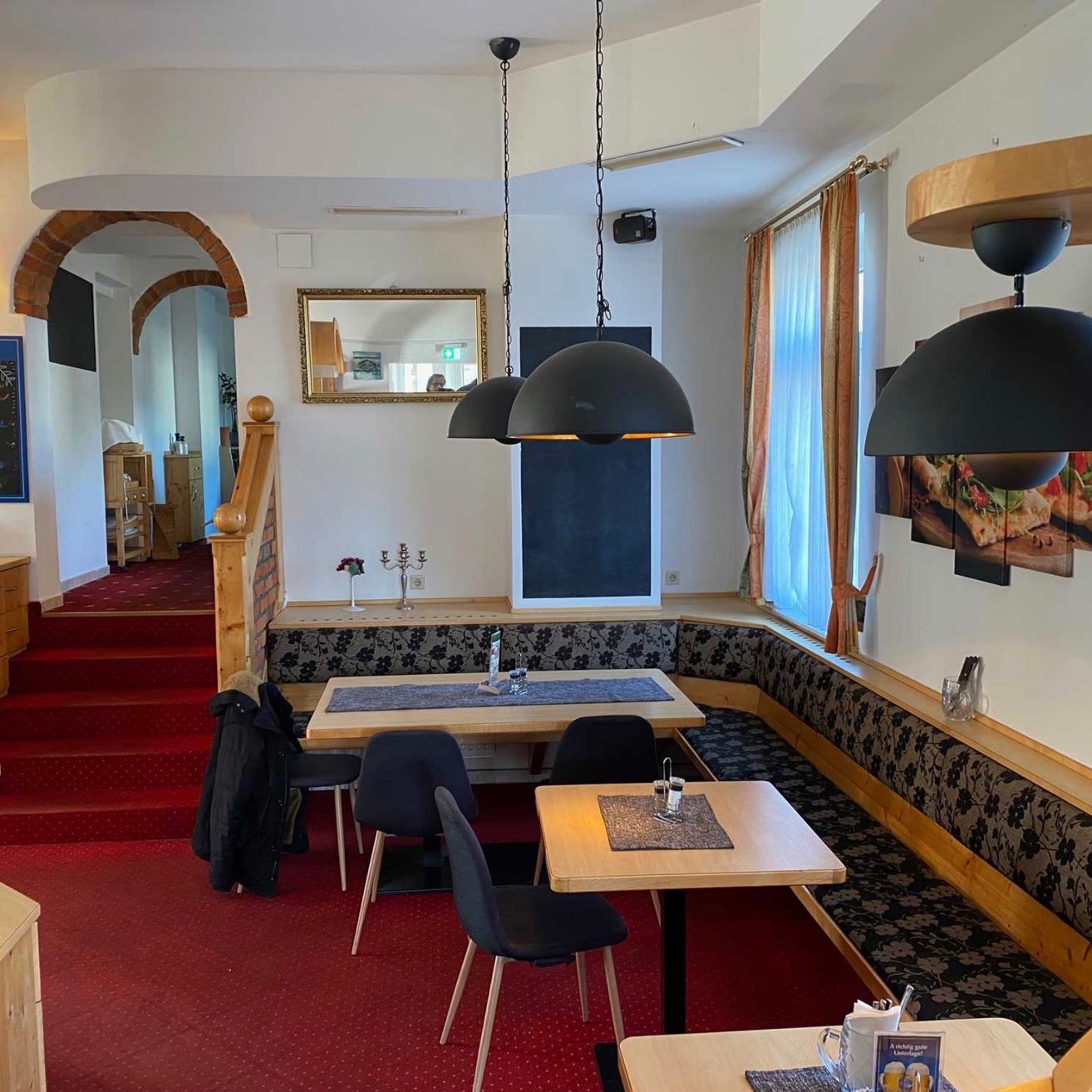 Ein Innenrestaurant mit einer Holzbank, rotem Teppich, Holztisch und Stühlen. Es hat schwarze Pendelleuchten und einen Spiegel an der Wand. Es gibt eine Tafel und einen Kerzenständer auf der Bank. An der Wand befinden sich auch einige Bilderrahmen.