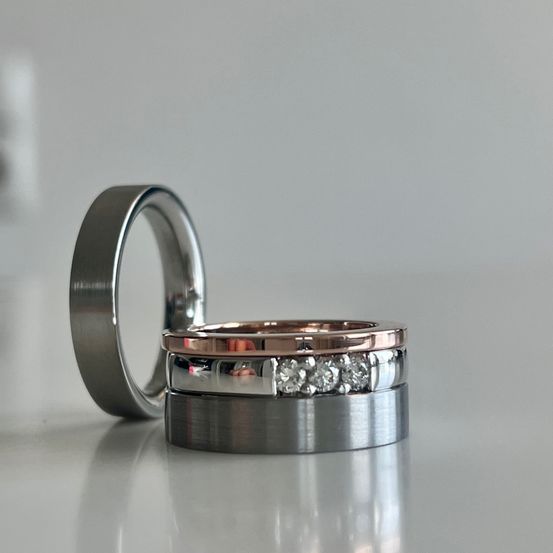 Drei Eheringe sind auf einer weißen Oberfläche gestapelt. Der obere Ring ist silber, der mittlere ist Roségold mit Diamanten und der untere Ring ist silber.