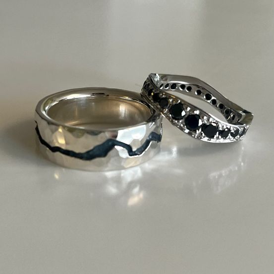 Zwei Silberringe sind auf einer weißen Oberfläche ausgestellt. Ein Ring hat eine gehämmerte Textur mit einer schwarzen Linie, und der andere ist mit schwarzen Steinen verziert.