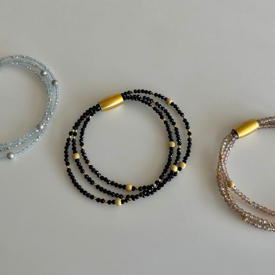 Drei Armbänder sind zu sehen. Das linke ist blau mit silbernen Perlen, das mittlere ist schwarz mit goldenen Perlen und das rechte ist rosa mit goldenen Perlen.