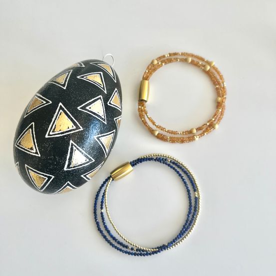 Ein dekorativer schwarz-goldener Eierschmuck mit geometrischem Muster, zwei Armbänder mit goldenen und blauen Perlen und ein golden-braunes Perlenarmband.