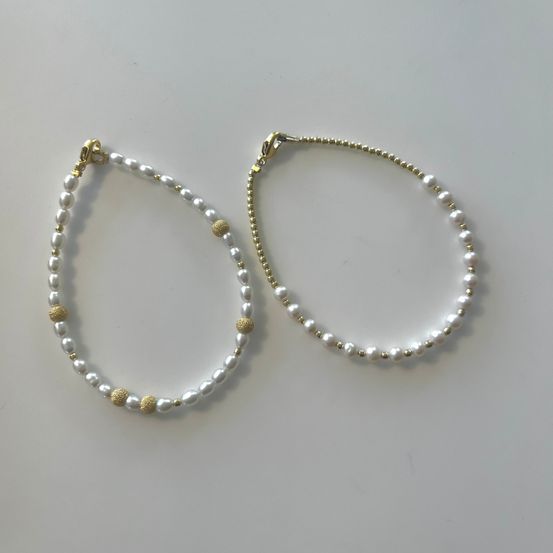 Zwei Perlenhalsketten mit goldenen Akzenten auf einer weißen Oberfläche ausgestellt. Eine Halskette hat goldene Perlen und die andere golden beschichtete Verschlüsse.