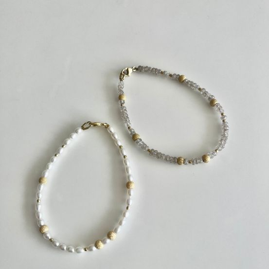 Zwei Perlenarmbänder, eines mit weißen Perlen und goldenen Akzenten, das andere mit klaren Kristallen und goldenen Akzenten, auf einem weißen Hintergrund gezeigt.