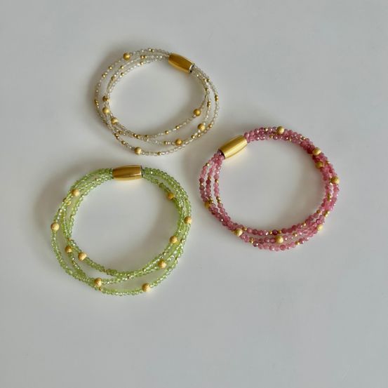Drei Armbänder mit goldenen Akzenten sind ausgestellt. Das oberste Armband ist weiß mit goldenen Perlen. Das mittlere ist grün. Das unterste Armband ist rosa.