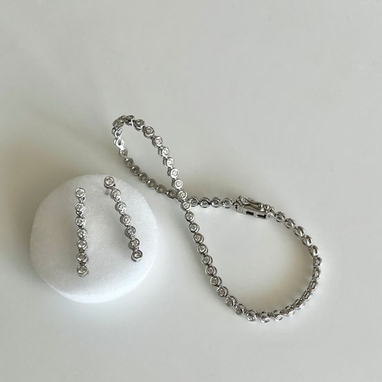 Eine Silberkette und passende Ohrringe sind auf einer weißen Oberfläche zu sehen. Die Kette besteht aus einer Gliederkette mit kleinen diamantartigen Perlen. Die Ohrringe sind lang und ebenfalls mit ähnlichen Perlen verziert.