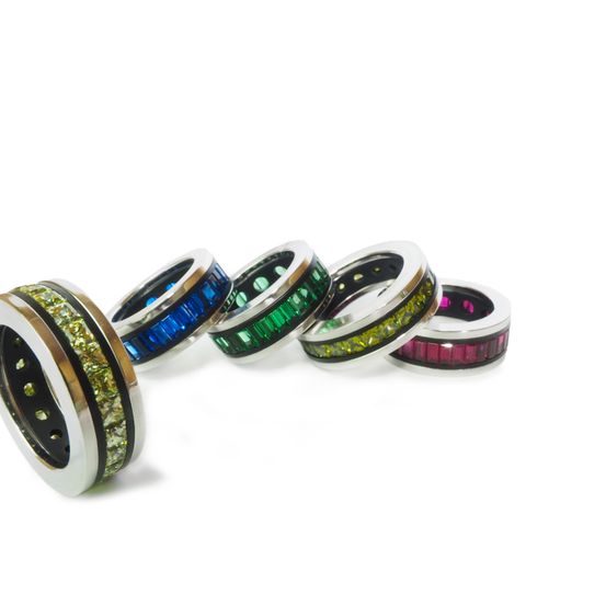 Fünf metallische Ringe mit farbigen Edelsteinen in Blau, Grün, Gelb und Rosa schmücken einen weißen Hintergrund.