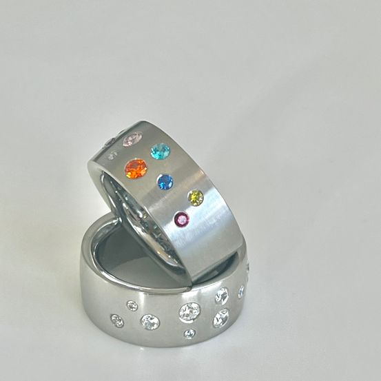 Ein silberner Ring mit mehrfarbigen Edelsteinen und kleinen Diamantstuds liegt auf einem weißen Untergrund auf einem anderen silbernen Ring.