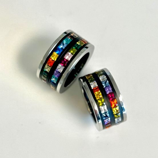 Zwei metallische Ringe, jeweils mit einem Regenbogen aus farbigen Steinen verziert, auf einer weißen Oberfläche gezeigt.