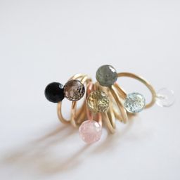 Ein goldener Ring mit mehreren kleinen Edelsteinperlen, darunter schwarze, rosa, grüne und durchsichtige Steine, vor weißem Hintergrund.