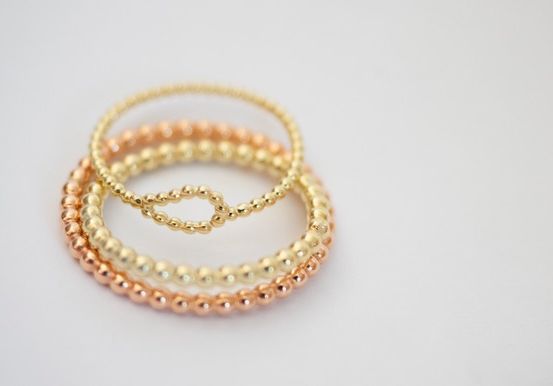 Drei gestapelte Armbänder mit goldenen und roségoldenen Perlen, auf einer weißen Oberfläche platziert.