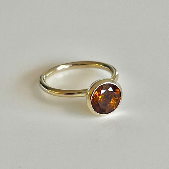 Ein goldener Ring mit einem runden orangefarbenen Edelstein, präsentiert auf einem weißen Hintergrund.