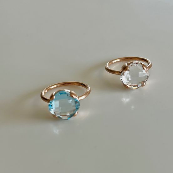 Zwei goldene Ringe mit blauen und klaren Kristallen sind auf einer weißen Oberfläche zu sehen. Die Ringe haben ein modernes, minimalistisches Design.