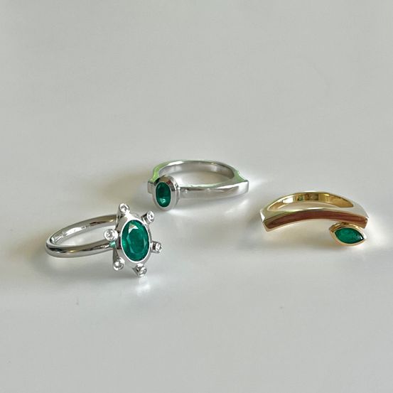 Drei Silber- und Goldringe mit Smaragd-Edelsteinen auf einer weißen Oberfläche. Jeder Ring hat ein einzigartiges Design.