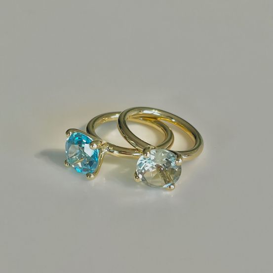 Zwei goldene Ringe mit blauen und weißen Edelsteinen sind auf einem weißen Hintergrund zu sehen. Die Ringe weisen kissenförmige Steine auf, einen blauen und einen weißen.