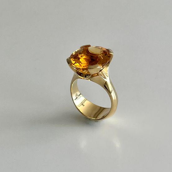 Ein goldener Ring mit einem großen, hellgelben Edelstein. Der Ring ist auf einem grauen Hintergrund zentriert.