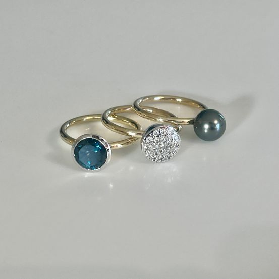 Drei Goldringe mit blauem Topas, Diamant und schwarzer Perle in der Mitte auf weißem Hintergrund gezeigt.