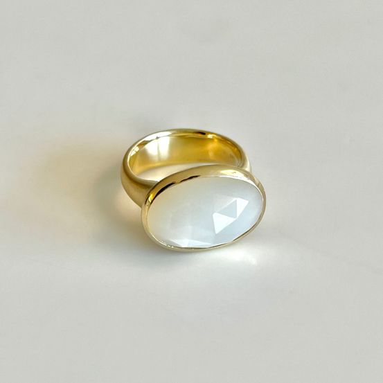 Ein goldener Ring mit einem weißen ovalen Stein in der Mitte, der auf einer weißen Oberfläche Licht reflektiert.