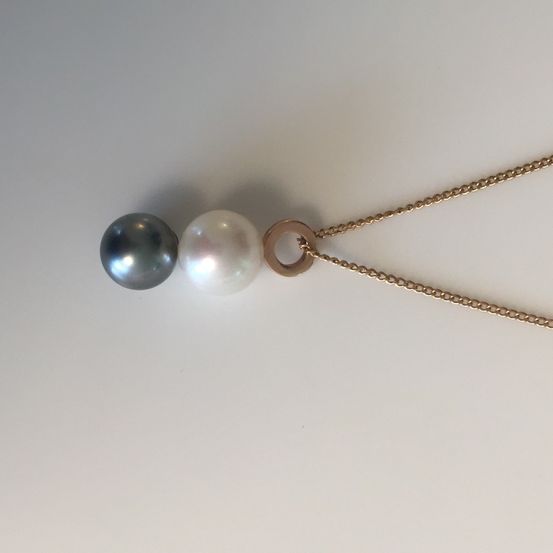 Eine Goldkette mit zwei Anhängern, eine schwarze Perle und eine weiße Perle, auf einer weißen Oberfläche.