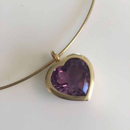 Ein goldener Herzanhänger mit einem lila Edelstein ruht auf einer goldenen Kette. Der Anhänger ist herzförmig und hat eine glänzende, polierte Oberfläche.