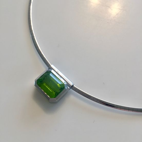 Eine silberne Halskette mit einem grün gefassten Edelstein-Anhänger mit quadratischem Schliff, auf einem weißen Hintergrund präsentiert.