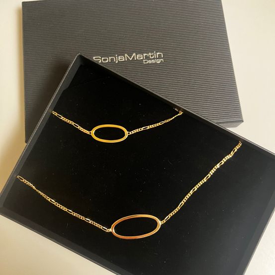 Zwei goldene Halsketten mit ovalen Anhängern in einer schwarzen Sonja Martin Design-Box. Die Anhänger sind kreisförmig und die Ketten sind gekettelt.