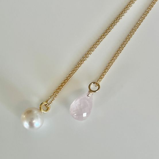 Zwei goldene Ketten mit Anhängern, eine mit einer Perle und die andere mit einem rosa Edelstein, auf einer weißen Oberfläche gezeigt.
