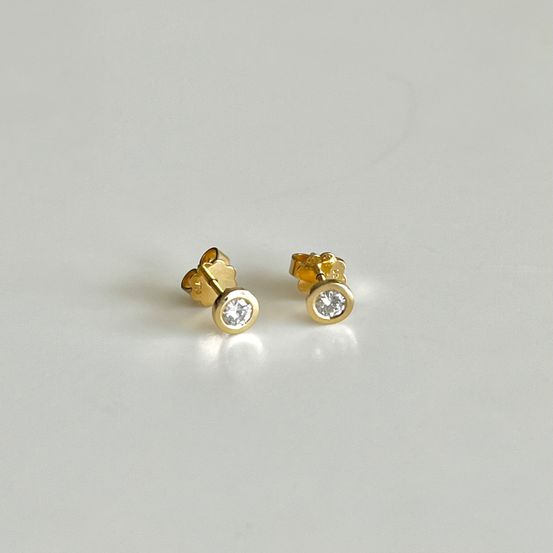 Zwei goldfarbene Ohrstecker mit einem kleinen Diamanten in der Mitte, auf einer weißen Oberfläche präsentiert.