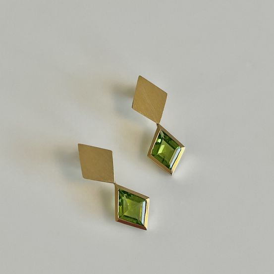 Zwei goldene Ohrringe mit grünen Edelsteinzentren, auf einer weißen Oberfläche präsentiert. Die Ohrringe haben eine Diamantform, bestehend aus Quadrat und Dreieck.
