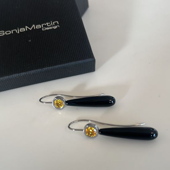 Zwei schwarze Ohrringe mit gelben Kristallen, präsentiert in einer Sonja Martin Design-Box auf einer weißen Oberfläche.