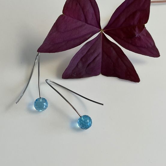 Zwei Ohrringe mit blauen Glasperlen, einer mit zwei Perlen, liegen neben einem violetten Blatt auf einer weißen Oberfläche.
