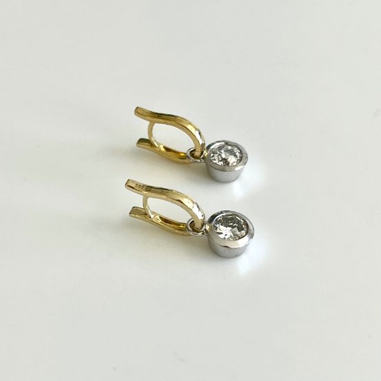 Zwei vergoldete Ohrringe mit einem kleinen Diamant-Mittelstück, auf einer weißen Oberfläche präsentiert.